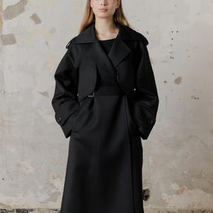 Op de afbeelding: Een zwarte, getailleerde trenchcoat met een riem en een lange rok. De jas heeft een kraag en lange mouwen.