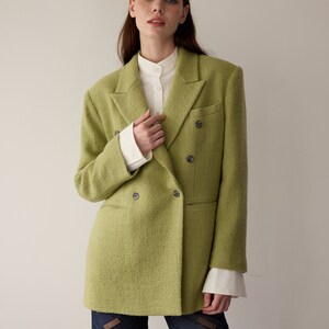 May include: A green double-breasted blazer with a textured weave. The blazer has a white collared shirt underneath and is worn with dark blue jeans.