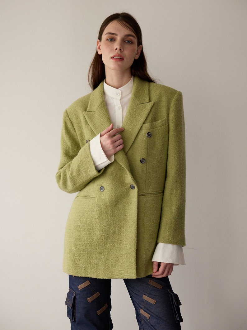 May include: A green double-breasted blazer with a textured weave. The blazer has a white collared shirt underneath and is worn with dark blue jeans.