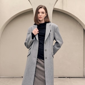 Op de afbeelding: Een vrouw draagt een lange, grijze wollen jas met een zwarte coltrui en een grijze rok. De jas heeft een klassiek ontwerp met knopen en zakken. De outfit heeft een neutraal kleurenpalet.