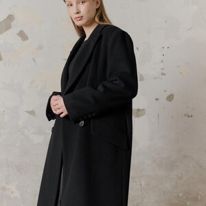 Op de afbeelding: Een vrouw draagt een lange zwarte jas met een dubbele rij knopen. De jas heeft een klassieke stijl en is gemaakt van een zachte, luxe stof.