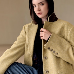 Op de afbeelding: Een vrouw draagt een lichtgele blazer met een bruine kraag en een zwarte hoodie. De blazer heeft vier knopen op de mouw en twee aan de voorkant. Ze draagt een zwarte top en een blauwe jeans.