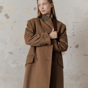 Op de afbeelding: Een bruine, dubbelrijdige wollen jas met een hoge kraag en lange mouwen. De jas wordt gedragen door een persoon met lang bruin haar.