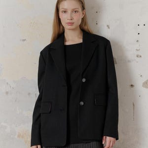 Op de afbeelding: Een zwart blazer met een enkele knoopsluiting en twee zakken. De blazer wordt gedragen over een zwart topje en een zwarte geplooide rok.