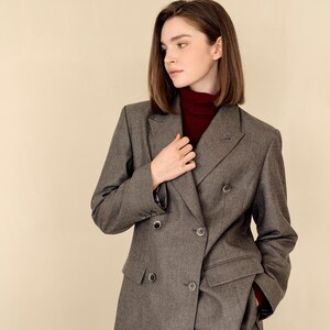 Op de afbeelding: Een grijze double-breasted blazer met bijpassende rok. De blazer heeft twee rijen knopen en een revers met inkeping. Daaronder wordt een bordeauxrode coltrui gedragen. De outfit is een klassieke, professionele stijl.