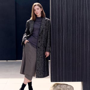 Op de afbeelding: Een vrouw draagt een grijze coltrui, rok en een lange zwart-wit gespikkelde jas. Ze staat naast een donkergrijze, getextureerde muur. Ze draagt zwarte enkellaarsjes.