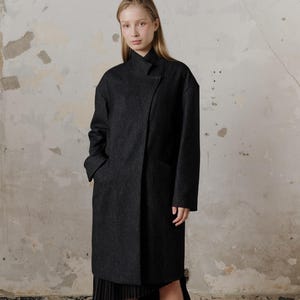 Op de afbeelding: Een lange, zwarte wollen jas met een hoge kraag en een enkele knoopsluiting. De jas heeft een licht asymmetrisch ontwerp en wordt gedragen over een zwarte geplooide rok.