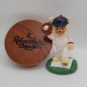 May include: A small, white, ceramic figurine of a bear wearing a baseball uniform with red and blue stripes. The bear is holding a baseball glove with a red baseball inside. The bear is standing on a green base with the words "A Robert Raikes Original TM" on a wooden base.