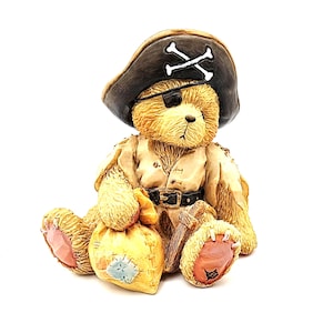 May include: A tan teddy bear figurine dressed as a pirate, wearing a black tricorn hat with a white skull and crossbones emblem, a black eye patch, and a cream-colored shirt with a black belt. The bear holds a yellow sack and a wooden sword.
