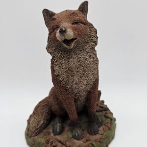 May include: A detailed figurine of a smiling fox, primarily brown with a lighter chest and face. The fox is seated on a base of leaves and earth tones. The figurine has a textured appearance, capturing the fox's fur and facial features.