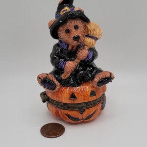May include: A ceramic Halloween trinket box featuring a witch bear riding a jack-o'-lantern. The bear is wearing a black witch hat with a purple trim and holding a broom.