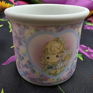 May include: A ceramic mug with a floral pattern and a heart-shaped graphic featuring a cartoon girl holding flowers. The mug has a white rim and a light pink and purple floral design.