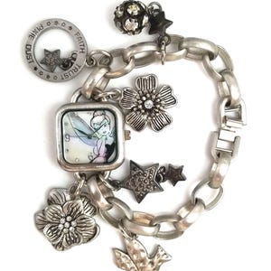 May include: A silver-tone charm bracelet featuring a square watch face with a Tinker Bell illustration. The bracelet includes various charms: two floral shapes with clear crystals, a bird, stars, a textured sphere, and a circular tag inscribed with Faith, Trust, and Pixie Dust.