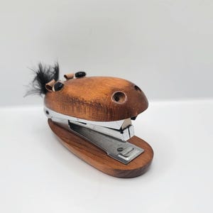 May include: A wooden stapler shaped like a horse head with a polished brown finish, featuring black bead eyes, a small tuft of black hair on top, and a silver metal stapling mechanism integrated into the base and upper jaw structure.