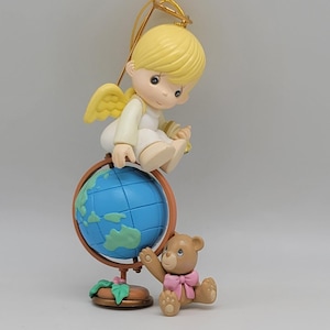May include: A white angel figurine with blonde hair and a gold halo sits on a blue globe. A brown teddy bear with a pink bow sits next to the globe.