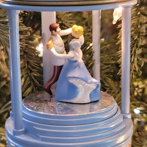 May include: A blue and gold musical ornament featuring Cinderella and Prince Charming dancing in a gazebo. The ornament has a gold crown on top and the words "Dreams Come True" on the bottom.