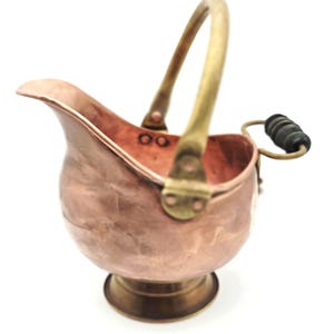May include: A small copper pitcher with a hammered texture, featuring a brass pedestal base, a brass swing handle, and a small brass side handle with a black wooden grip. The interior rim shows two small circular markings.
