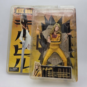 May include: Two action figures from the movie "Kill Bill" are displayed in their original packaging. One figure is of "The Bride" in a yellow jumpsuit, holding a sword. The packaging features the movie title and character details.