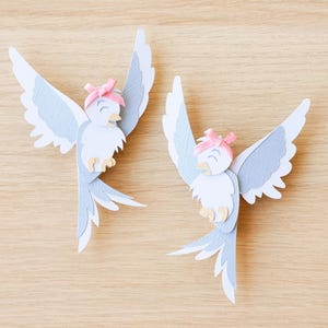 Puede incluir: Dos decoraciones de pájaros de papel con las alas extendidas. Los pájaros tienen cuerpos blancos, alas azul claro y lazos rosas. Están sobre un fondo de madera marrón claro. Los pájaros están hechos con recortes de papel detallados.