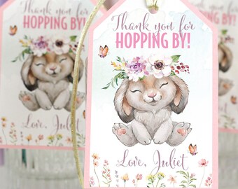 Könnte beinhalten: Rechteckige Geschenkanhänger mit rosa Rand und einer Aquarellillustration eines braunen Hasen mit Blumenkranz. Der Text lautet Thank you for HOPPING BY! und Love, Juliet. Jeder Anhänger ist mit einer goldenen Schnur an einer Glasflasche befestigt.