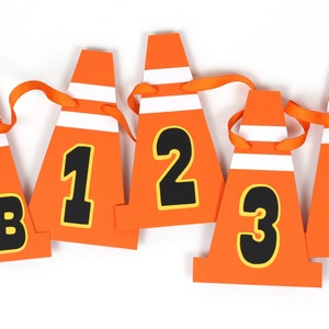 May include: Orange and white construction cone shaped paper cutouts with black numbers 1, 2, 3, 4 and the letters NB. The cutouts are strung together with orange ribbon.