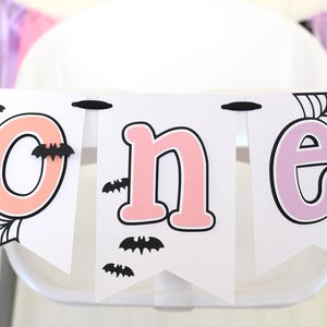 May include: A decorative banner featuring three white pennants strung on a black ribbon. The letters O, N, and E are printed in peach, pink, and light purple. The design includes black spider web illustrations and small black bat cutouts.