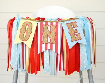 May include: A high chair banner featuring three pennants with the word ONE in gold glitter letters. The pennants are yellow, red and white striped, and light blue, accented with hanging ribbons in red, gold, and light blue polka dots.