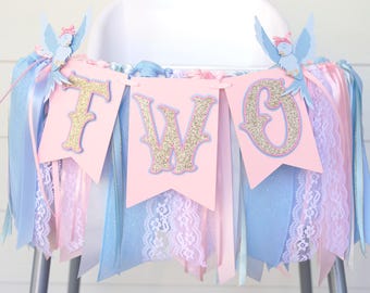 May include: A high chair banner featuring three light pink pennants with gold glitter letters spelling TWO. The banner is decorated with blue bird cutouts with pink bows and a fringe of light blue, pink, and white ribbons and lace.