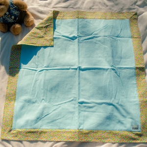 May include: A light blue baby blanket with a small checkered pattern. The blanket has a floral patterned border in shades of green, orange, and yellow. A teddy bear wearing a blue floral vest is in the upper left corner.