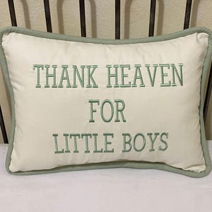 May include: Cream-colored rectangular pillow with the embroidered text "THANK HEAVEN FOR LITTLE BOYS" in green. The pillow has a green border and is placed on a white surface.