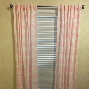 May include: A pair of pink and white patterned curtains hang in front of a window with white blinds. The curtains are made of a lightweight fabric and have a subtle sheen.