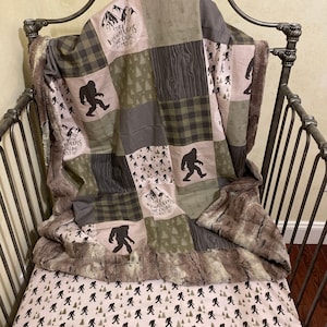 May include: A baby crib with a green and brown patchwork quilt featuring a Bigfoot design and the text "The Mountains Are Calling But I Must Go". The crib sheet is white with a repeating pattern of Bigfoot silhouettes.