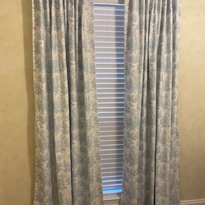 May include: A pair of blue and white patterned curtains hang in front of a window with white blinds. The curtains have a toile pattern.