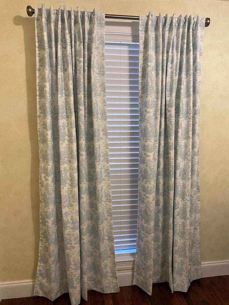 May include: A pair of blue and white patterned curtains hang in front of a window with white blinds. The curtains have a toile pattern.