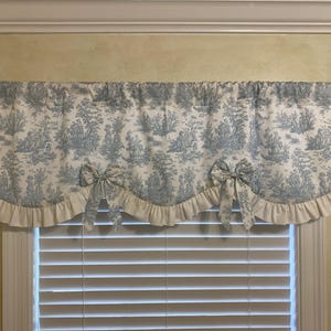 May include: A blue and white toile valance with a ruffled edge and two decorative bows. The fabric features a pastoral scene. The valance is mounted on a dark brown curtain rod above a window with white blinds.