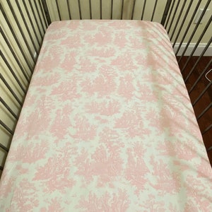 Op de afbeelding: Een roze en wit gestreept hoeslaken voor een babybedje met een toile-patroon.