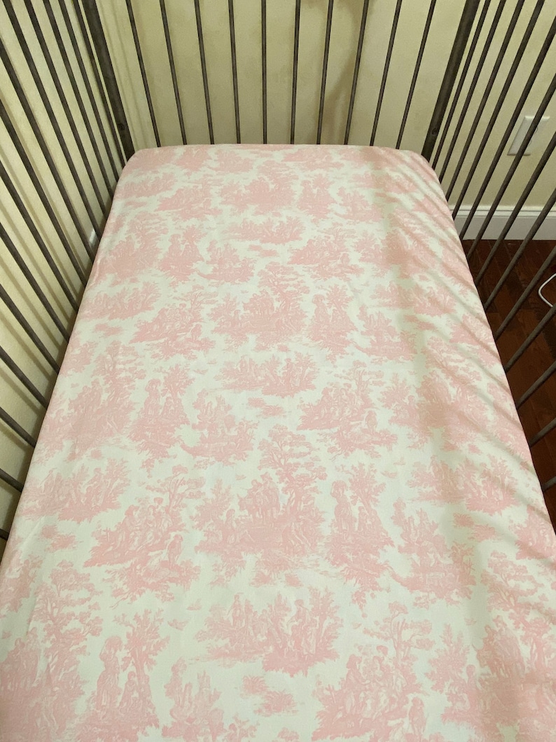 Op de afbeelding: Een roze en wit gestreept hoeslaken voor een babybedje met een toile-patroon.