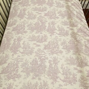 Op de afbeelding: Een wit babybeddengoed met een lavendelkleurig toile-patroon met een herhalend ontwerp van bomen, bloemen en mensen.