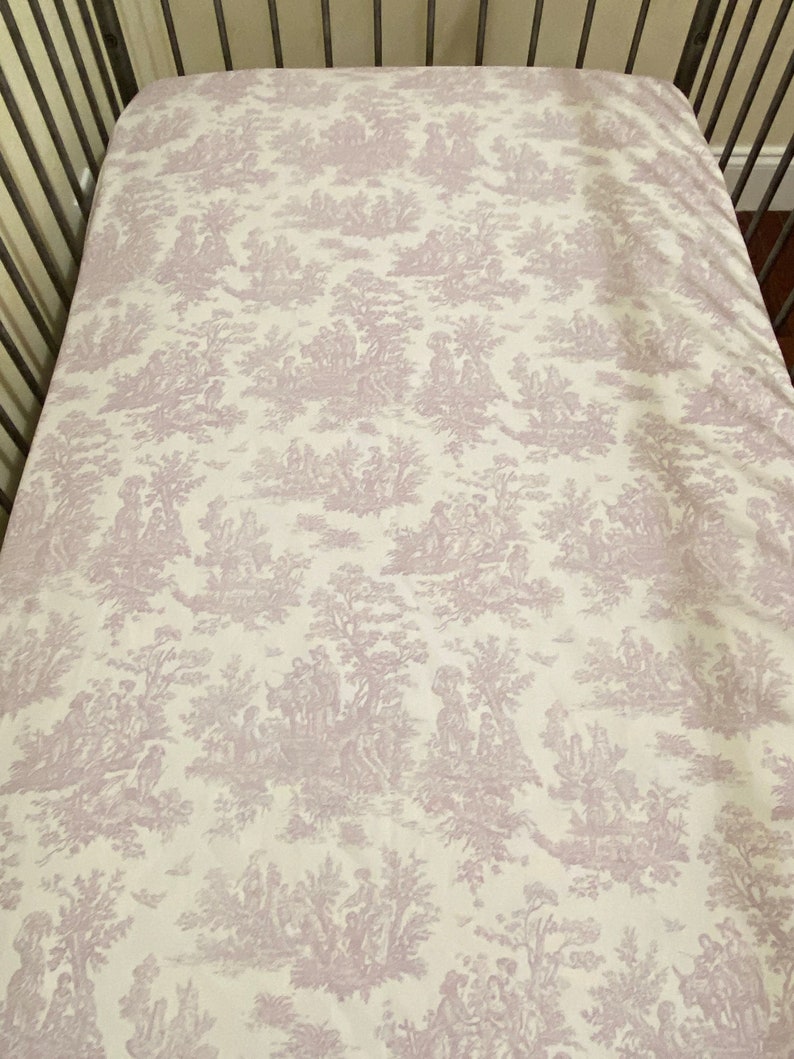 Op de afbeelding: Een wit babybeddengoed met een lavendelkleurig toile-patroon met een herhalend ontwerp van bomen, bloemen en mensen.