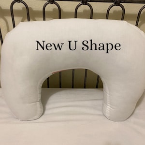 May include: A white, U-shaped pillow with the text "New U Shape" printed on the front. The pillow is on a white sheet, and the background includes a metal headboard.