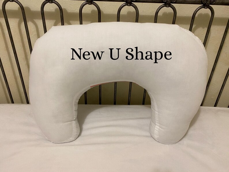 May include: A white, U-shaped pillow with the text "New U Shape" printed on the front. The pillow is on a white sheet, and the background includes a metal headboard.