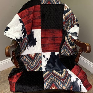 May include: A patchwork quilt with a variety of patterns, including red and white woodgrain, black, and a geometric pattern in shades of blue, red, and beige. The quilt is folded and draped over a wooden rocking chair.