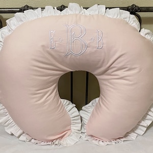 May include: A light pink nursing pillow with white ruffled trim. The pillow has a crescent shape with a central opening. The center of the pillow features the embroidered letters "E B B" in white.