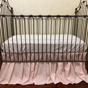 Pu&ograve; includere: Una culla in ferro battuto con un materasso bianco e una gonna a quadretti rosa e bianchi. La culla ha una testiera e una pediera decorative. La gonna della culla &egrave; arricciata e ha un bordo a balze.