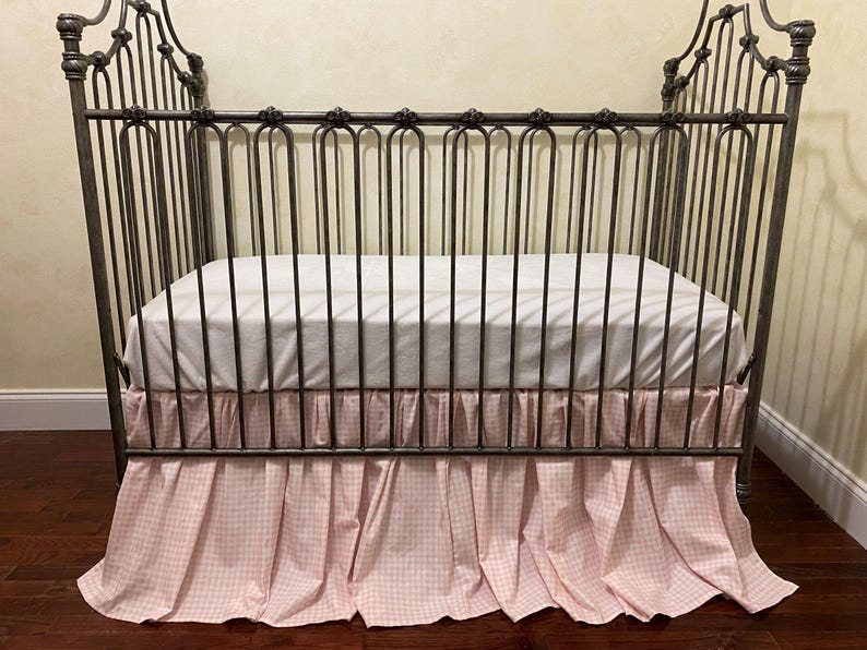 Pu&ograve; includere: Una culla in ferro battuto con un materasso bianco e una gonna a quadretti rosa e bianchi. La culla ha una testiera e una pediera decorative. La gonna della culla &egrave; arricciata e ha un bordo a balze.