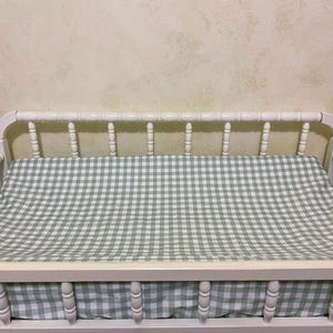 May include: A white wooden changing table with a green and white gingham patterned changing pad. The changing table has a decorative spindle design on the headboard and footboard.