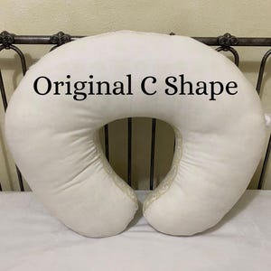 May include: Cream-colored, C-shaped nursing pillow with the text "Original C Shape" in black. The pillow is made of a soft, plush material and has a decorative trim along the edges. The pillow is on a white surface.