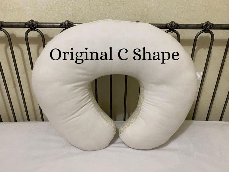 May include: Cream-colored, C-shaped nursing pillow with the text "Original C Shape" in black. The pillow is made of a soft, plush material and has a decorative trim along the edges. The pillow is on a white surface.