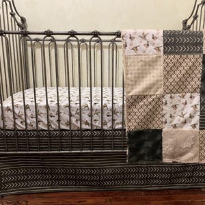 May include: A metal crib with a dark finish, featuring a fitted sheet with a duck pattern and a matching bed skirt with a white chevron design. A patchwork quilt is draped over the side, displaying squares of duck print, tan plaid, dark faux fur, and geometric patterns.