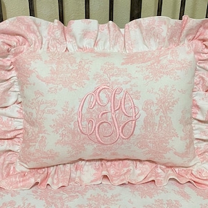 May include: A pink and white pillow with a ruffled edge and a monogram in the center. The pillow has a toile pattern with pink flowers and trees.